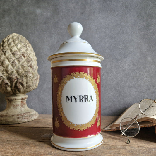 Decorative jar labeled 'MYRRA' on a wooden surface with a textured gray wall background.