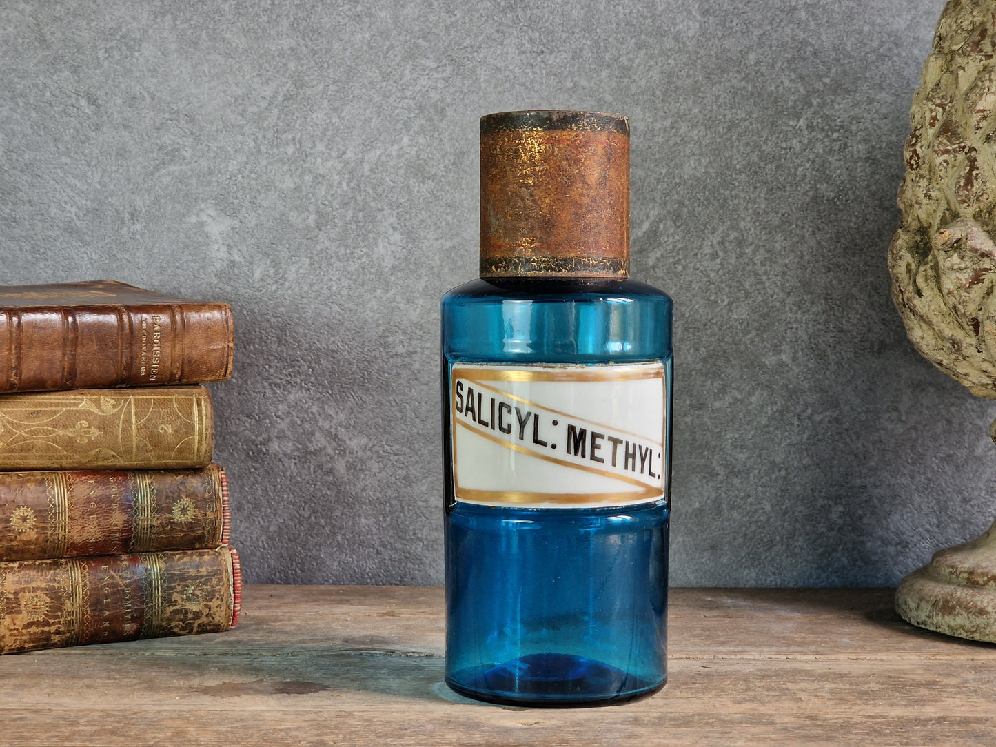 An antique cobalt blue glass bottle with a rustic copper-toned cap and a white label reading 'SALICYL: METHYL', displayed on a wooden surface against a gray wall with old books and a stone bust in the background. 