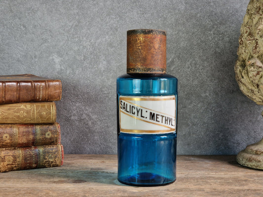 An antique cobalt blue glass bottle with a rustic copper-toned cap and a white label reading 'SALICYL: METHYL', displayed on a wooden surface against a gray wall with old books and a stone bust in the background. 