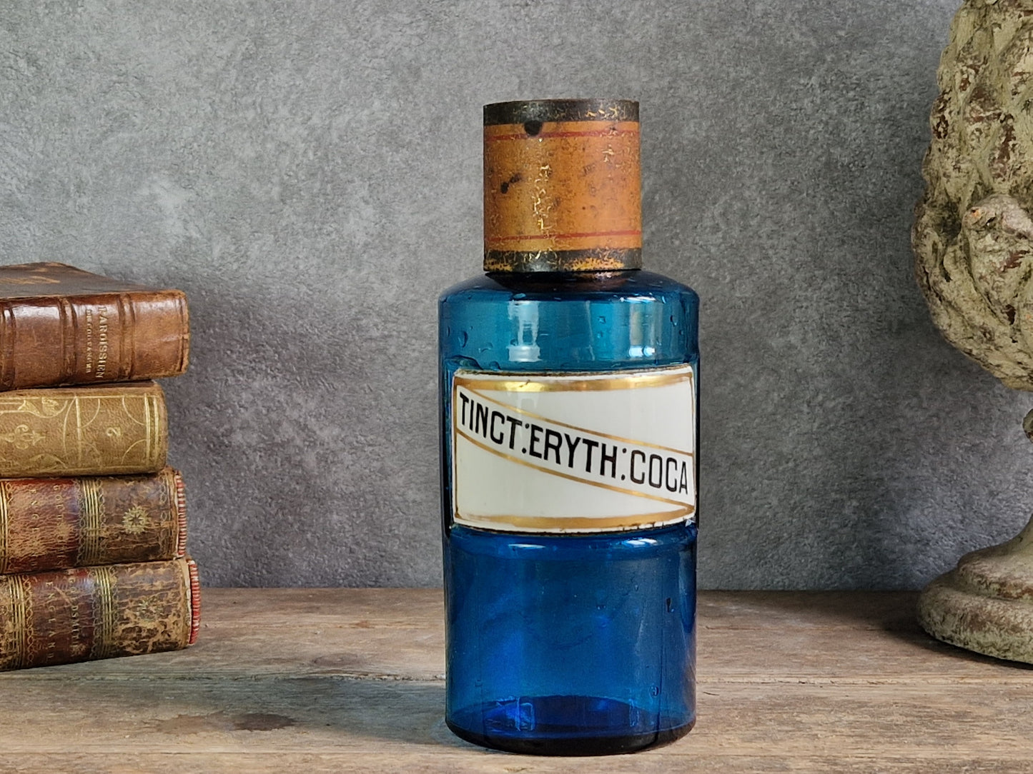 An antique cobalt blue glass bottle with a white label reading 'TINCT ERYth: COCA', displayed on a wooden surface against a gray wall with old books and a stone bust in the background.