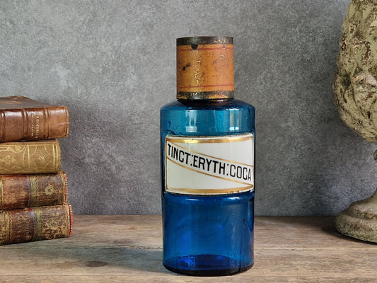 An antique cobalt blue glass bottle with a white label reading 'TINCT ERYth: COCA', displayed on a wooden surface against a gray wall with old books and a stone bust in the background.