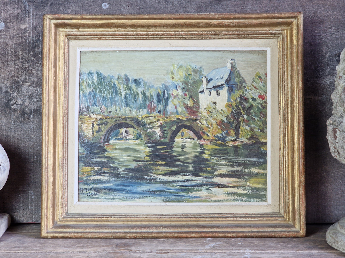 A framed landscape painting depicting a stone bridge over water with trees and a house, set in a distressed gold frame against a rustic wooden background.