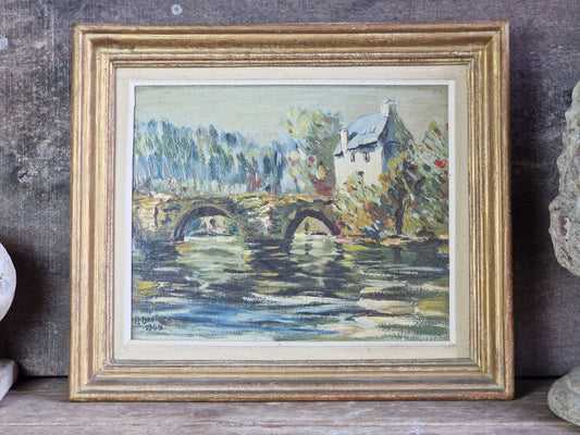 A framed landscape painting depicting a stone bridge over water with trees and a house, set in a distressed gold frame against a rustic wooden background.